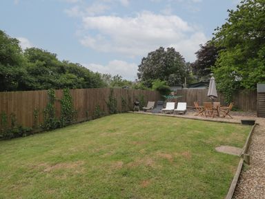 A garden with lawn chairs a round wooden table with an umbrella a barbecue and wooden fencing at Pigsfoot Lodge in Ash Thomas near Halberton Devon