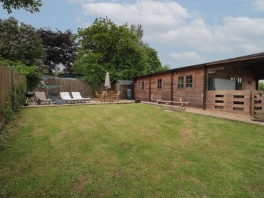 A backyard with lawn chairs a wooden table with umbrella a barbecue and a wooden building at Pigsfoot Lodge in Ash Thomas near Halberton Devon
