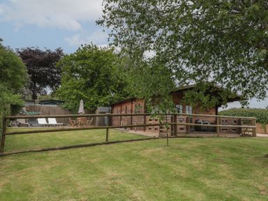 A wooden cabin with a fenced lawn and outdoor furniture including lounge chairs and a table with an umbrella at Pigsfoot Lodge in Ash Thomas near Halberton Devon