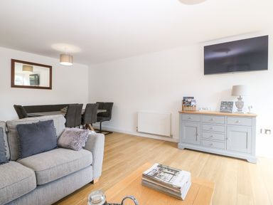 A living room with a grey sofa and cushions a wooden coffee table with magazines a grey sideboard with a lamp and decor a wall-mounted TV a dining area with six chairs at 3 Pensyflog Barns in Porthmadog
