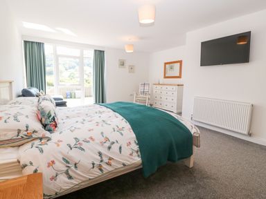 A bedroom with a bed a dresser a rocking chair a wall-mounted tv and large windows with curtains at 3 Pensyflog Barns in Porthmadog