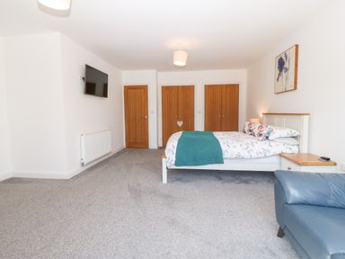 A bedroom with a bed covered in floral bedding and a teal throw a blue armchair a wall-mounted TV wooden doors and grey carpet at 3 Pensyflog Barns in Porthmadog