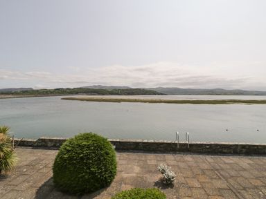 A waterfront view with a stone patio, bushes, a low stone wall, water, and hills in the background at 25 South Snowdon Wharf in Porthmadog