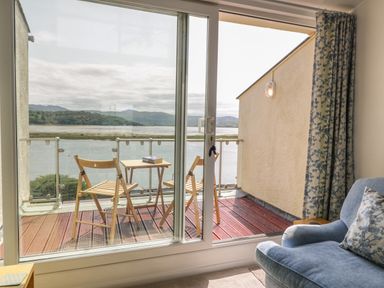 A wooden balcony with two folding chairs and a small table with a book overlooking a body of water seen through a sliding glass door next to a light blue armchair at 25 South Snowdon Wharf in Porthmadog