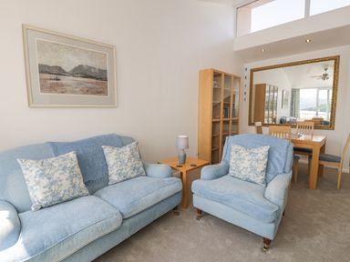 A living room with blue sofa and chair a wooden table with lamp a bookshelf and dining table with chairs at 25 South Snowdon Wharf in Porthmadog