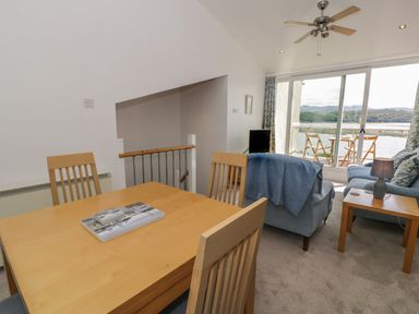 A dining area with wooden table and chairs next to a living room with blue sofas and a side table with lamp by sliding glass doors and balcony at 25 South Snowdon Wharf in Porthmadog
