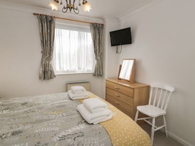 A bedroom with a bed covered in patterned bedding and folded towels next to a window with curtains a wooden dresser with a mirror and a wall-mounted TV and a white chair at 25 South Snowdon Wharf in Porthmadog