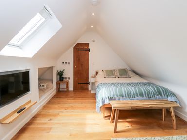 An attic bedroom with a bed a wooden bench a skylight window a wall-mounted television a small table with a plant and a wooden door at The Rookery in Fulbrook near Burford