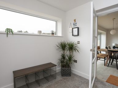 A room with a wooden bench with wire storage compartments a large plant in a pot small plants on a window ledge and a door opening to a dining area at Ysgol Fawr Big School in Benllech