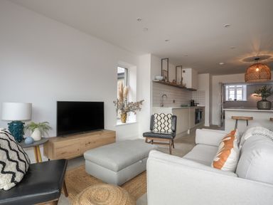 A living room with a grey sofa, black chairs, a wooden TV stand, and an open kitchen with stools at Ysgol Fawr (Big School) in Benllech