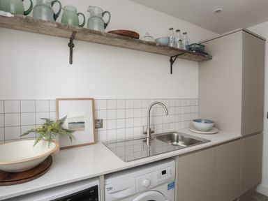 A kitchen countertop with a sink washing machine and wall shelf with pitchers and bowls at Ysgol Fawr Big School in Benllech