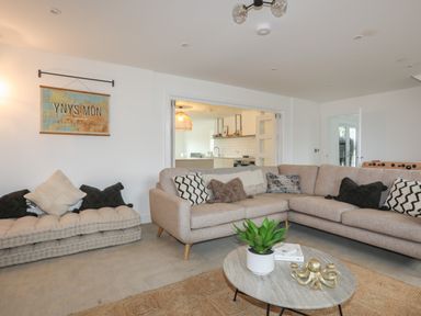 A living room with a beige sectional sofa and cushions a low cushioned bench with pillows a round coffee table with a plant and decoration a view into a kitchen at Ysgol Fawr Big School in Benllech