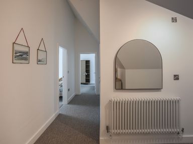 A hallway with two framed pictures on the left wall a radiator and an arched mirror on the right wall and two open doorways at Ysgol Fawr Big School in Benllech