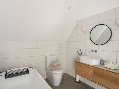 A bathroom with a bathtub a toilet with towels a wooden vanity with a sink a round mirror and wall tiles at Ysgol Fawr Big School in Benllech