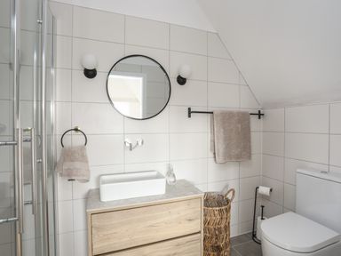 A bathroom with a wooden vanity with a rectangular white sink a round mirror above a toilet and towels at Ysgol Fawr Big School in Benllech