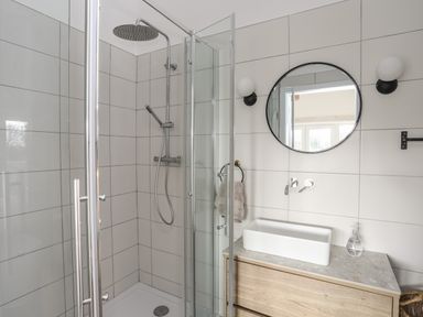 A bathroom with a glass shower enclosure a rain shower head a rectangular sink on a wooden vanity and a round mirror at Ysgol Fawr Big School in Benllech
