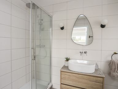 A bathroom with a glass shower enclosure a wooden vanity with a white sink an oval mirror and two wall lights at Ysgol Fawr Big School in Benllech