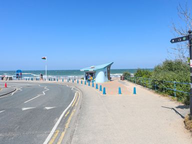A seaside road with a small blue kiosk near the beach and a flagpole with the Welsh flag at Ysgol Fawr Big School in Benllech