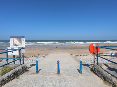 A concrete path leading to a sandy beach with waves and people near the shoreline at Ysgol Fawr Big School in Benllech