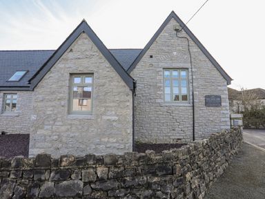 A stone building with pointed roofs and windows behind a stone wall at Ysgol Fawr Big School in Benllech