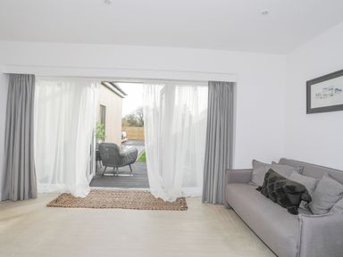 A living room with a gray sofa and cushions next to glass doors with curtains leading to an outdoor seating area at Ysgol Fawr Big School in Benllech