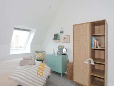 A bedroom with a bed a window a green chest of drawers a chair a wicker basket and a wooden bookcase at Ysgol Fawr Big School in Benllech