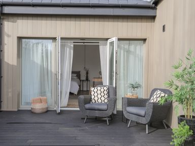 An outdoor patio with two woven armchairs with cushions a small side table with a plant and a potted plant near open double glass doors revealing a bedroom at Ysgol Fawr Big School in Benllech