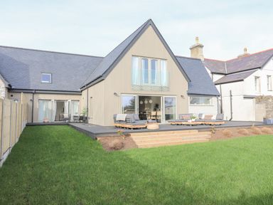 A modern house with a sloped roof and outdoor seating on a wooden deck with a green lawn at Ysgol Fawr Big School in Benllech