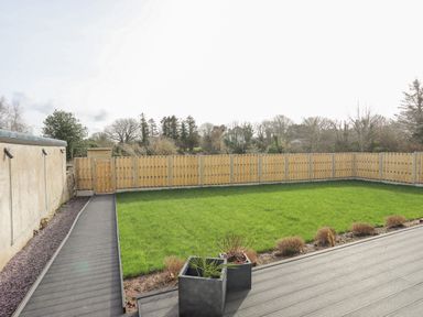 A garden with green grass and shrubs enclosed by a wooden fence with a paved pathway and planters at Ysgol Fawr Big School in Benllech
