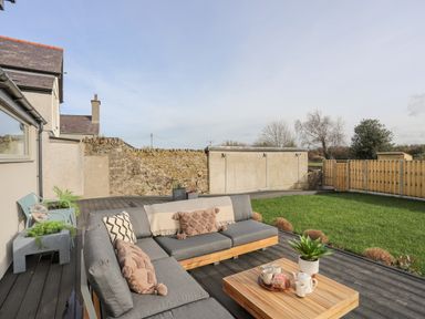 An outdoor patio with a grey sectional sofa, a wooden coffee table, potted plants, grass lawn, and a stone wall at Ysgol Fawr Big School in Benllech