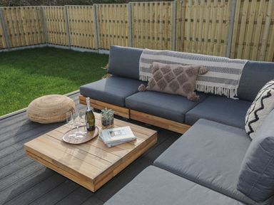 An outdoor seating area with a grey cushioned sofa a wooden coffee table with a bottle and glasses and a woven floor cushion at Ysgol Fawr Big School in Benllech