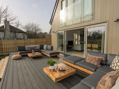 An outdoor patio with grey cushioned wooden sofas and two low wooden tables with cups and plants at Ysgol Fawr Big School in Benllech