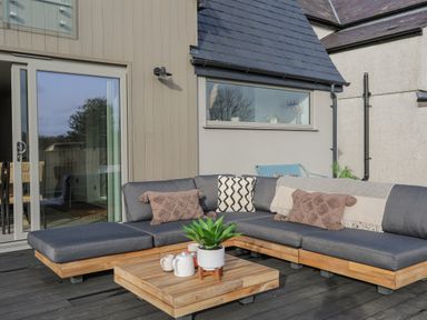 An outdoor patio with a wooden sectional sofa with gray cushions and pillows a wooden coffee table with a plant a teapot and cups on it and a glass sliding door at Ysgol Fawr Big School in Benllech