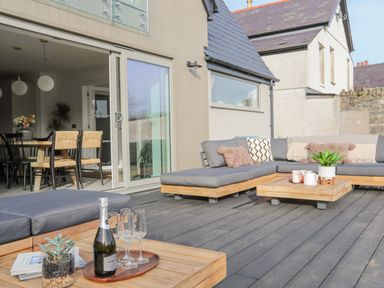 An outdoor patio with wooden deck furniture including a sectional sofa and tables with a bottle and glasses next to a dining area inside a house at Ysgol Fawr Big School in Benllech