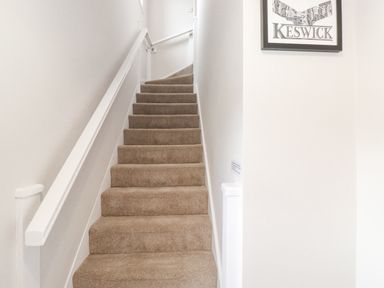A carpeted staircase with white walls and a handrail with a framed Keswick print on the wall at 1 Millbank in Keswick