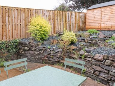 An outdoor patio area with a green metal table and chairs next to a stone retaining wall with plants and a wooden fence at 1 Millbank in Keswick