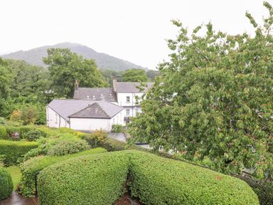 A view of white buildings with gray roofs behind green hedges and trees with hills in the background at 1 Millbank in Keswick