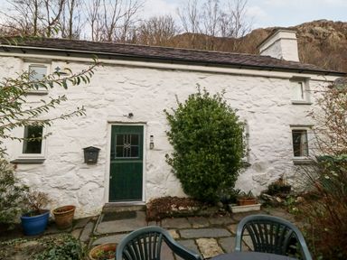 A stone cottage with a green door and windows a bush and potted plants outside with a plastic patio table and chairs at Is Y Graig in Beddgelert