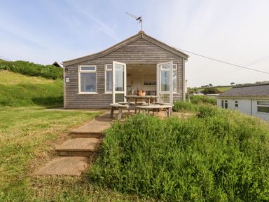A wooden chalet with open doors and a picnic table outside on a grassy area at Siesta Chalet in Eype Mouth Chalet Park