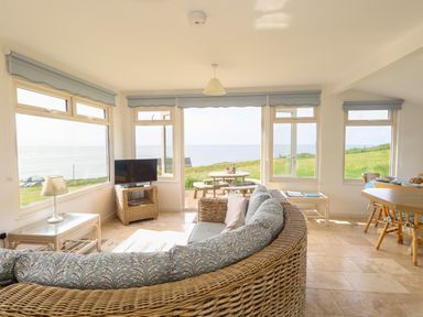 A living room with wicker furniture a TV and large windows overlooking a grassy area and sea at Siesta Chalet in Eype Mouth Chalet Park