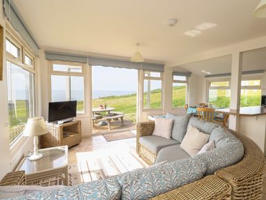 A living room with wicker furniture a television and dining area with outdoor view at Siesta Chalet in Eype Mouth Chalet Park