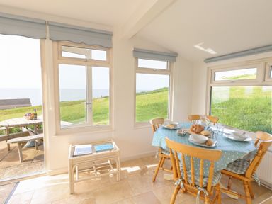 A dining area with a round table covered with a blue polka dot tablecloth and wooden chairs near large windows with a view of grass and sea at Siesta Chalet in Eype Mouth Chalet Park