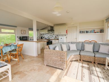 An open plan kitchen and living area with a wicker sectional sofa a dining table and chairs kitchen cabinets and tiled floor at Siesta Chalet in Eype Mouth Chalet Park