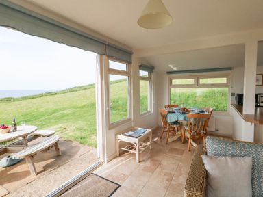 A dining area with wooden chairs and a table set near windows overlooking a grassy hill and a picnic table outside at Siesta Chalet in Eype Mouth Chalet Park
