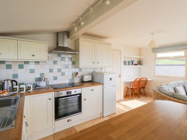 A kitchen with oven, fridge, toaster, kettle, mugs and a small dining area with wooden chairs and a window at Siesta Chalet in Eype Mouth Chalet Park