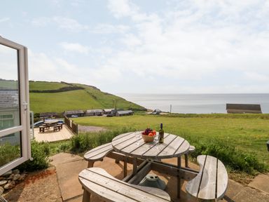 An outdoor wooden picnic table with fruit bowl wine bottle and glasses overlooking green hills and the sea at Siesta Chalet in Eype Mouth Chalet Park