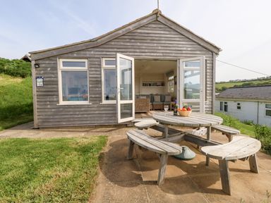 A wooden chalet with open double doors leading to a living area and a round wooden picnic table with benches outside at Siesta Chalet in Eype Mouth Chalet Park