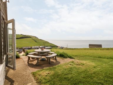 A patio with a round wooden picnic table and benches overlooking a grassy hill and sea at Siesta Chalet in Eype Mouth Chalet Park