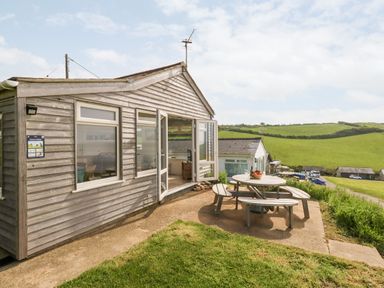 An exterior view of a wooden chalet with an open door and a round picnic table with benches on a concrete patio at Siesta Chalet in Eype Mouth Chalet Park