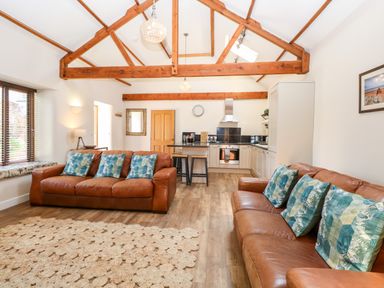 An open-plan living area with brown leather sofas, blue patterned cushions, a kitchen with white cabinets, wooden beams on the ceiling, and a wooden floor at The Barnhouse Felmingham near North Walsham